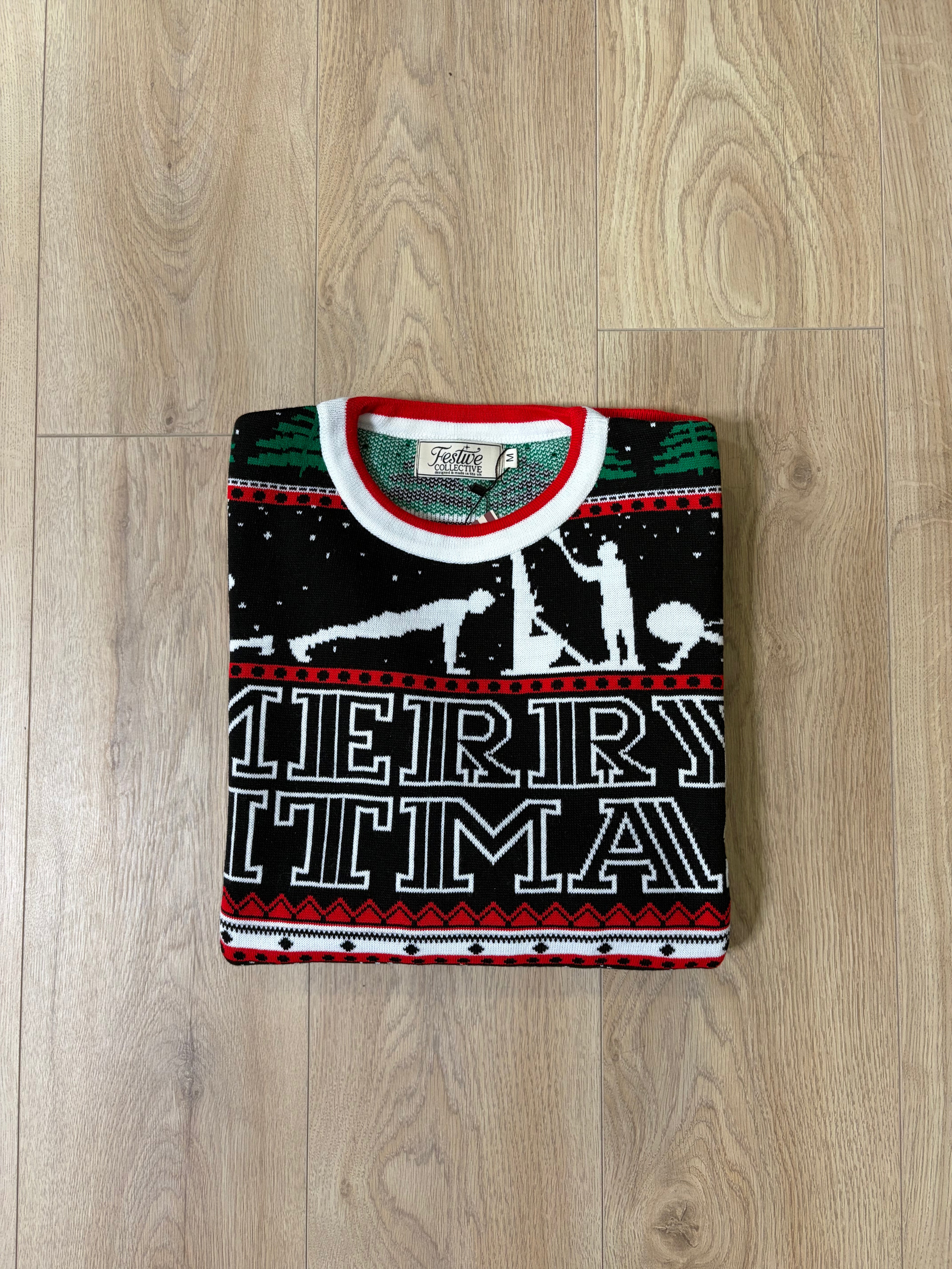 A folded black Christmas sweater with a festive pattern and 'MERRY CHRISTMAS' text, displayed flat on a light wood floor.
