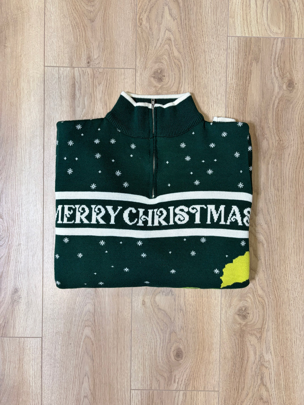 A dark green Christmas half-zip sweater with white snowflakes, a white 'MERRY CHRISTMAS' text band, and a yellow accent on the bottom right, laid flat on a light wood floor.