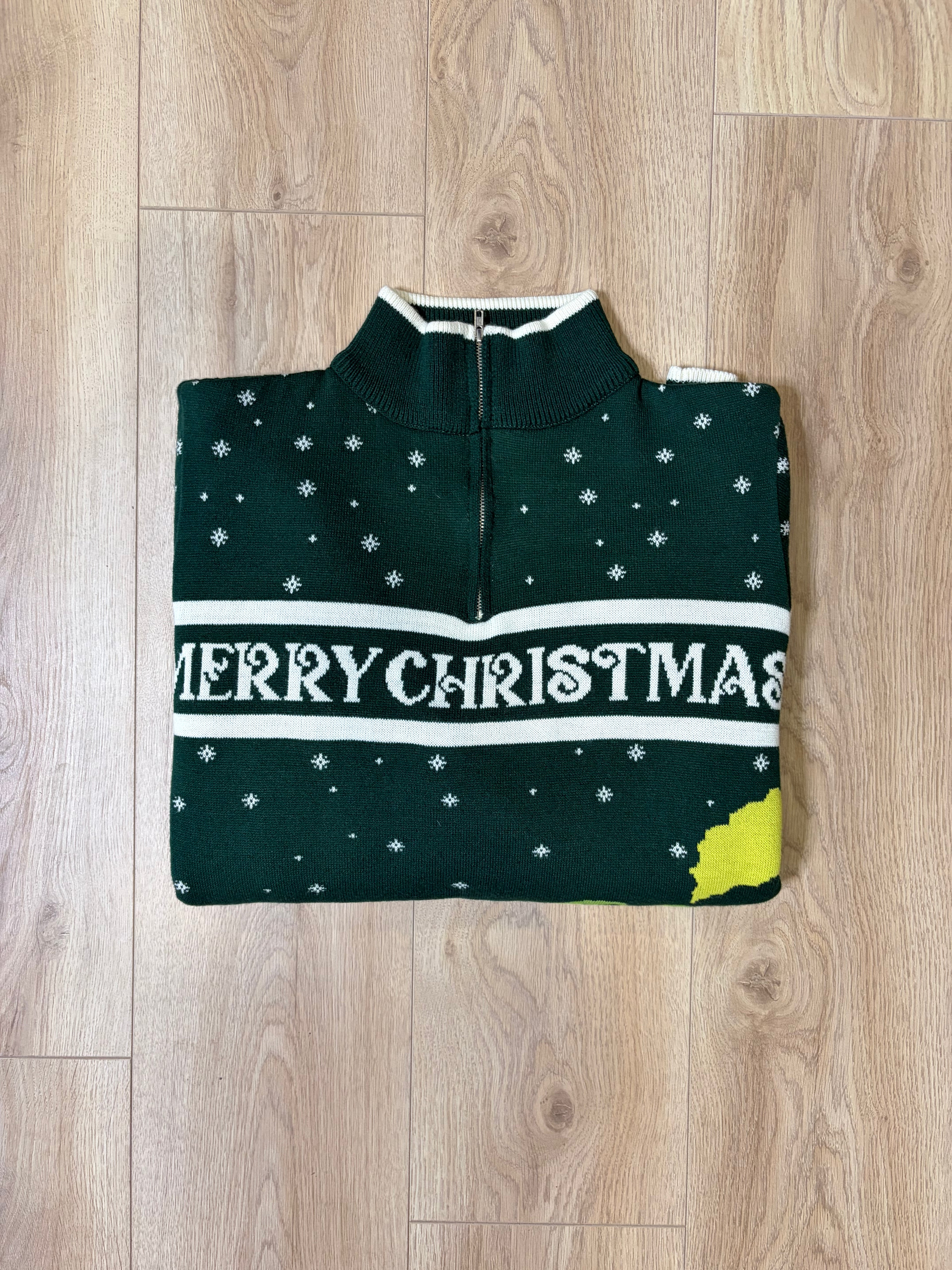 A dark green Christmas half-zip sweater with white snowflakes, a white 'MERRY CHRISTMAS' text band, and a yellow accent on the bottom right, laid flat on a light wood floor.
