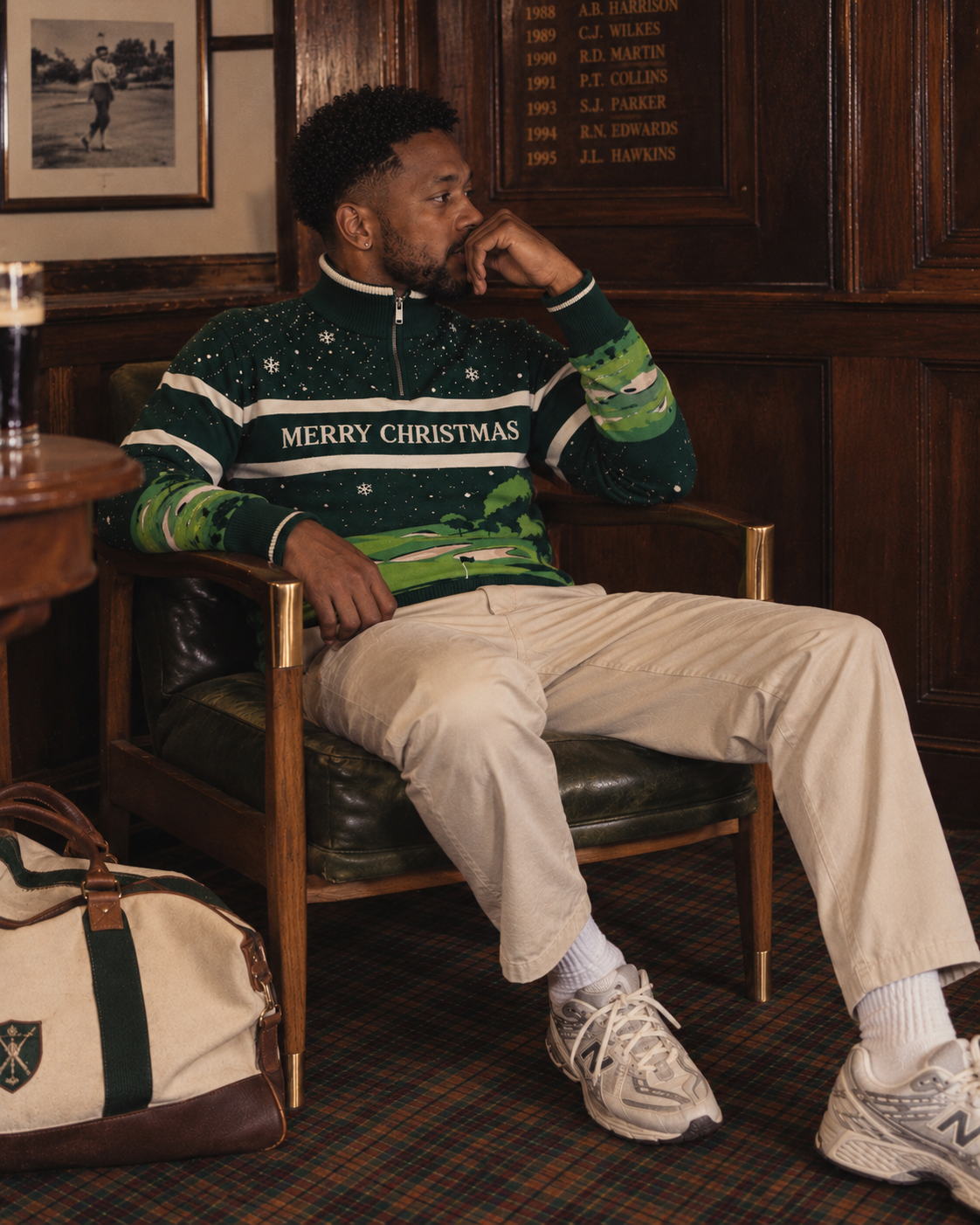 A man sits in a leather armchair in a wood-paneled room wearing a green Christmas quarter-zip sweater with 'MERRY CHRISTMAS' across the chest, cream trousers, white socks, and New Balance sneakers. A matching bag rests on the floor nearby.