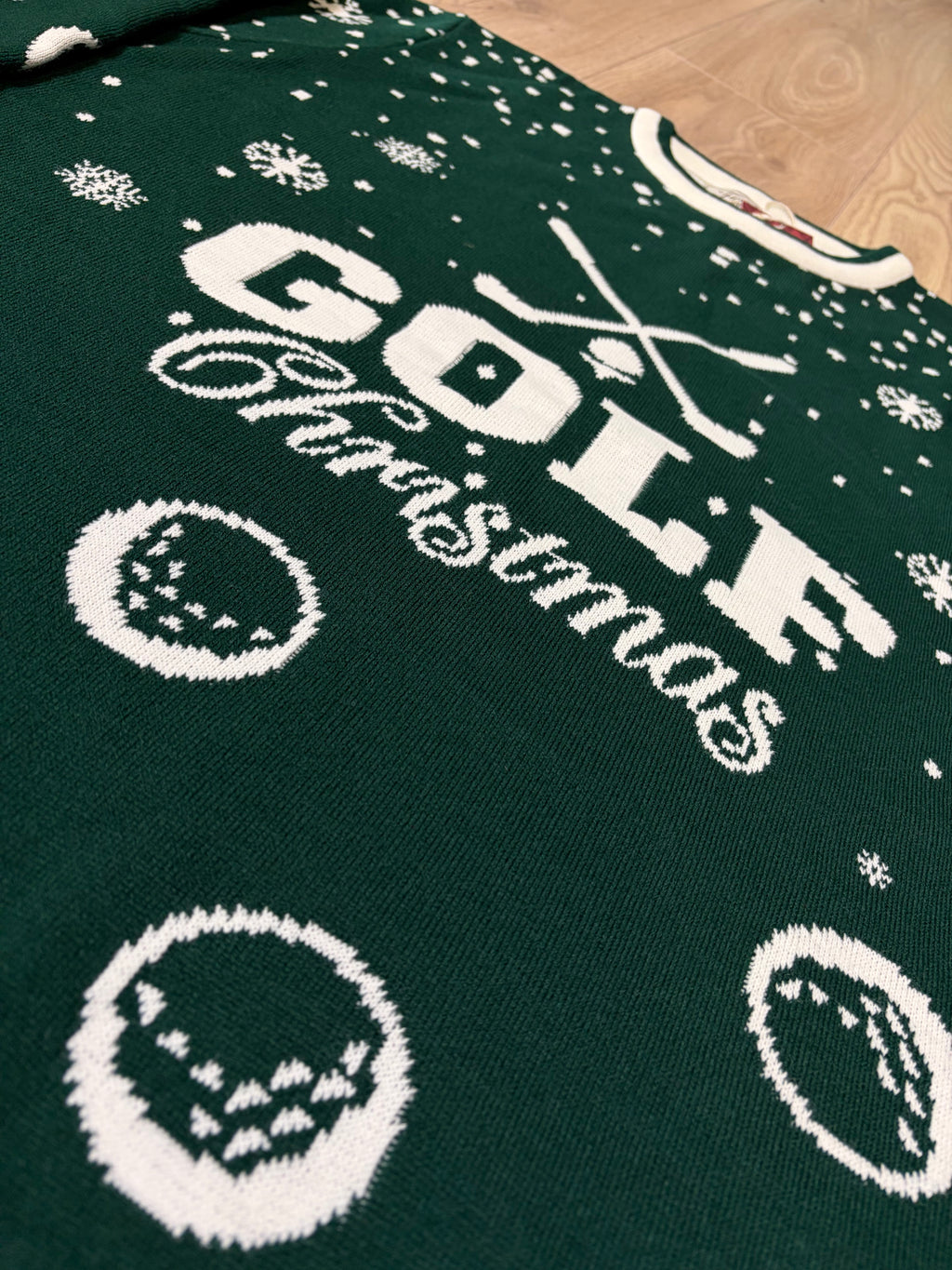 A close-up of a dark green knitted Christmas sweater with white knit graphics reading 'GOLF Christmas,' plus golf balls, snowflakes, and crossed clubs, laid on a light wood floor.