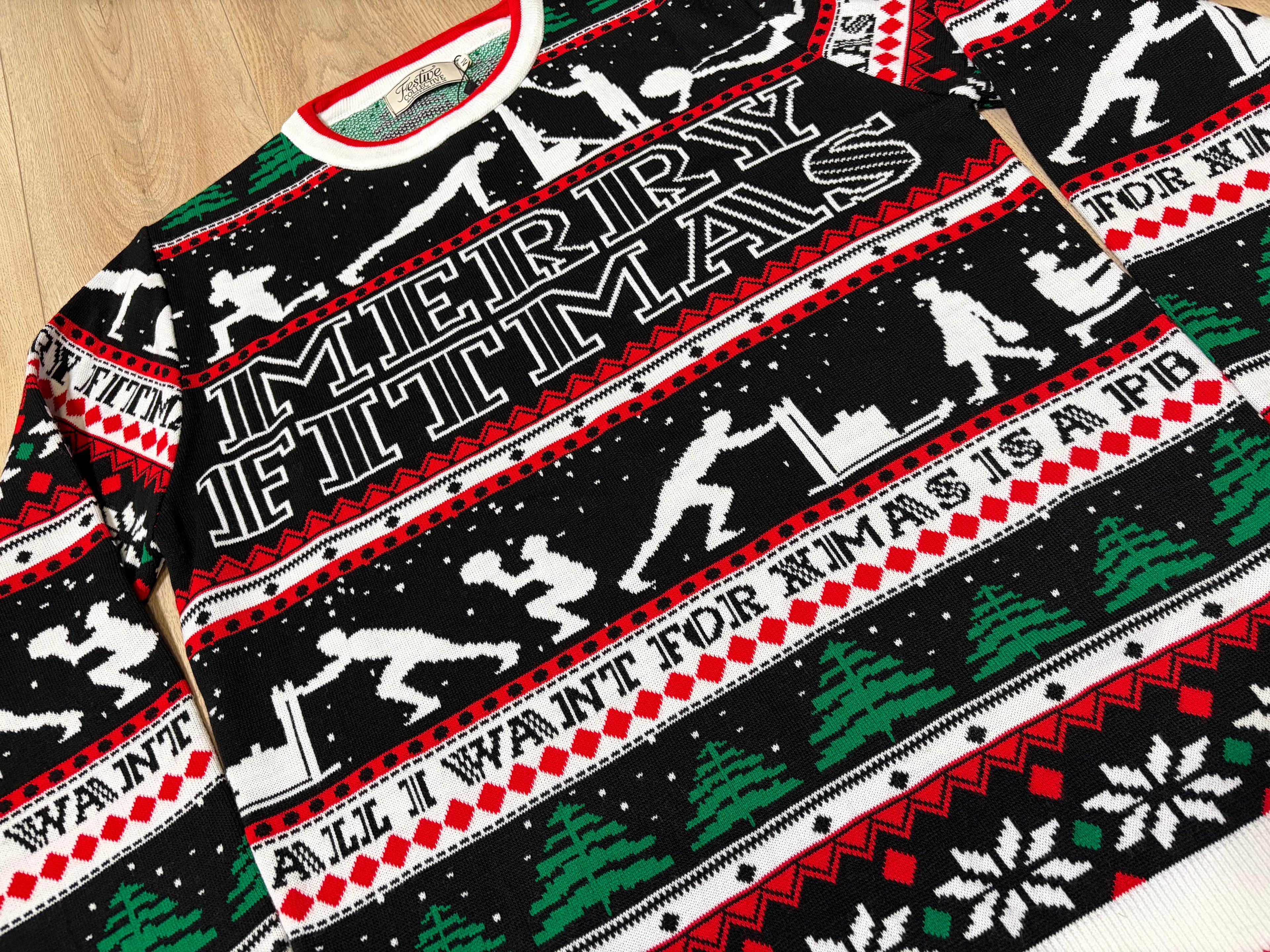 A close-up of a black Christmas sweater featuring a bold white and red knit pattern with the words 'MERRY CHRISTMAS' and 'ALL I WANT FOR XMAS IS A PB', along with silhouettes of people doing gym activities and festive tree motifs.