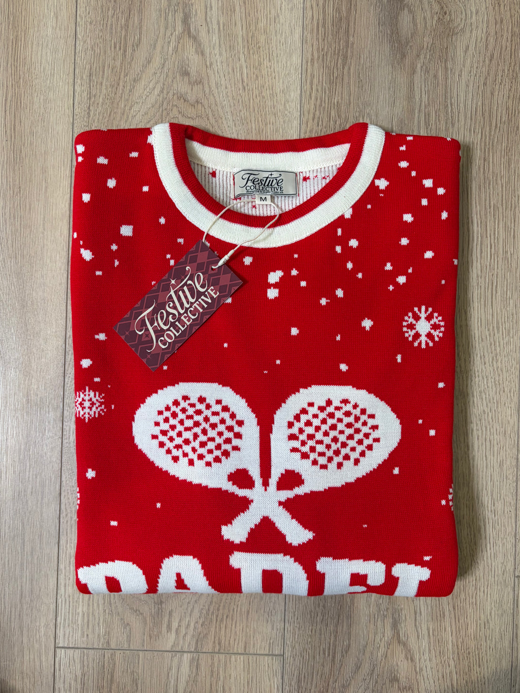 A folded red knit sweater with white snowflake and padel graphics, featuring a visible 'Festive Collection' hangtag, laid flat on a light wood floor.