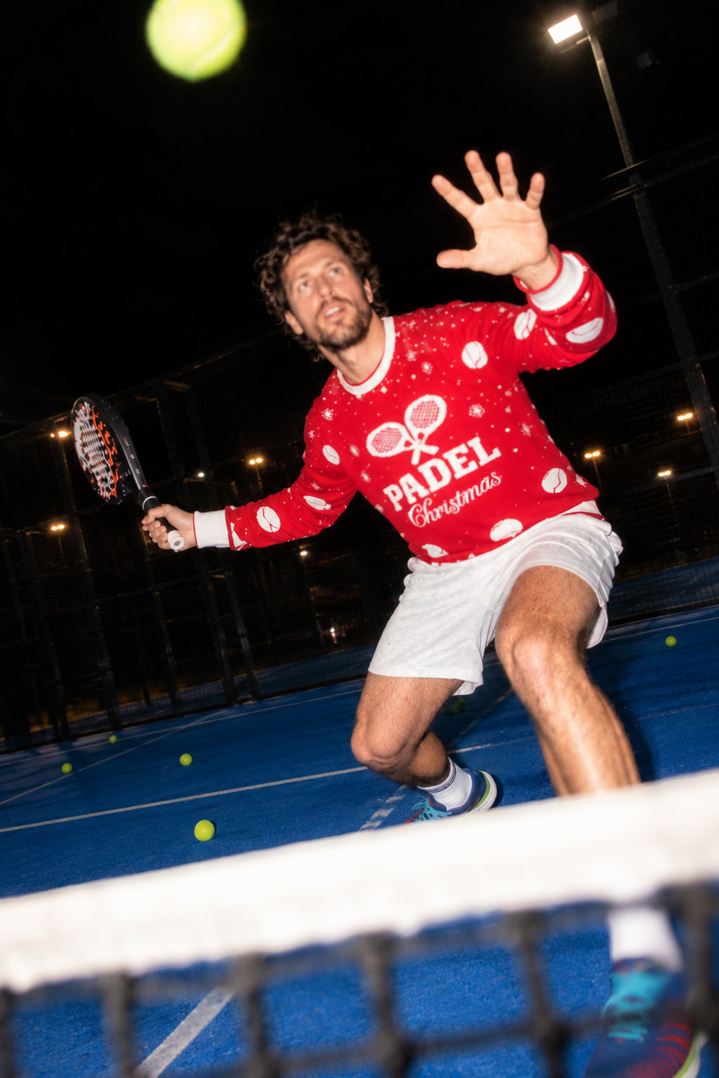 A man on a blue tennis court at night wearing a red and white Christmas-themed 'PADEL Christmas' sweater, white shorts, and sneakers, reaching for a tennis ball while holding a paddle.