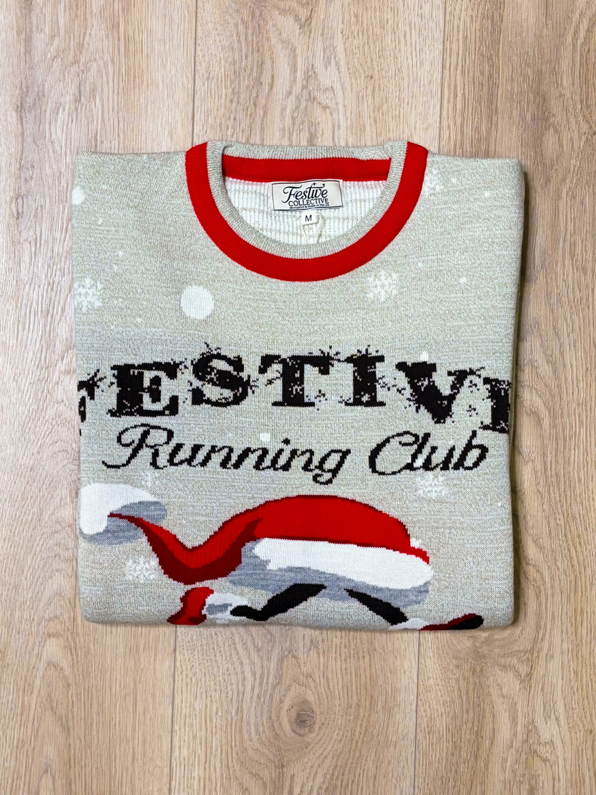 A folded light gray knit sweater with a red crew neck collar, featuring a festive graphic that reads 'FESTIV eRunning Club' with a red and white Santa hat and snowflake details, laid flat on a light wood floor.