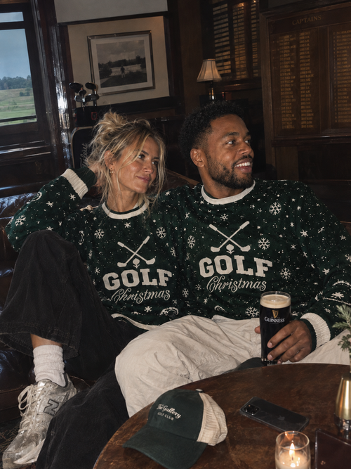 A man and a woman sitting together in a dimly lit pub wearing matching dark green Christmas sweaters with white snowflake and golf club graphics and the words 'GOLF Christmas'. The man holds a pint of Guinness.