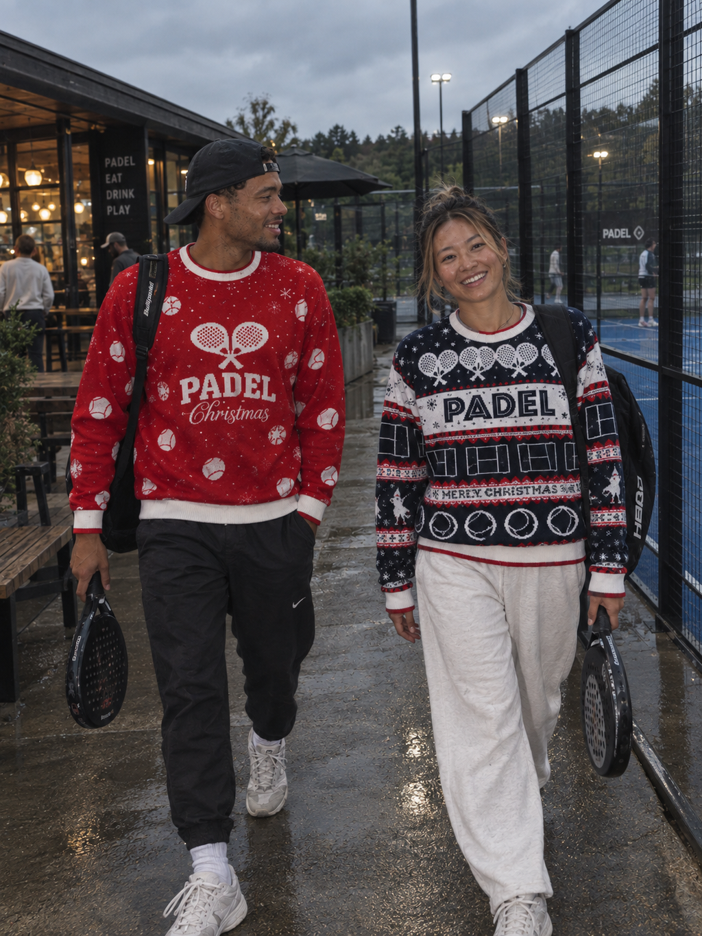 A man and a woman walking side-by-side on a wet paved path near a padelcourt, wearing festive Christmas sweaters. The man's sweater is red with white patterns and 'PADEL Christmas' text, and the woman's sweater is navy with red and white patterns and 'PADEL CHRISTMAS' text.