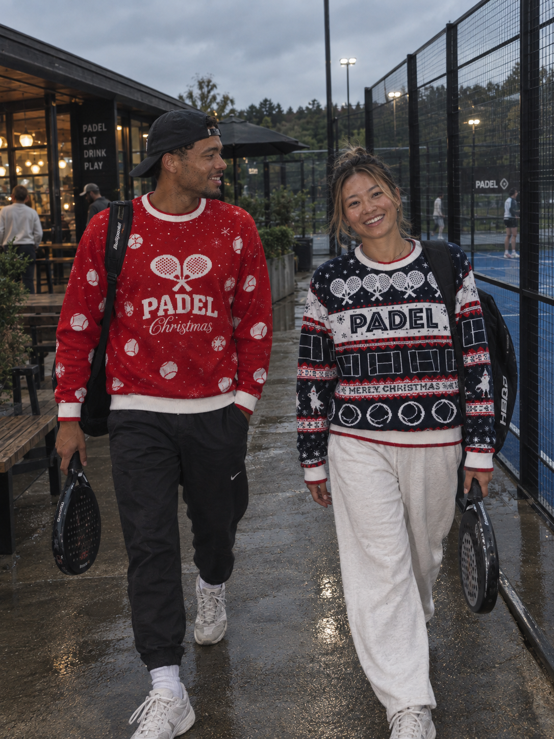 A man and a woman walking side-by-side on a wet paved path near a padelcourt, wearing festive Christmas sweaters. The man's sweater is red with white patterns and 'PADEL Christmas' text, and the woman's sweater is navy with red and white patterns and 'PADEL CHRISTMAS' text.