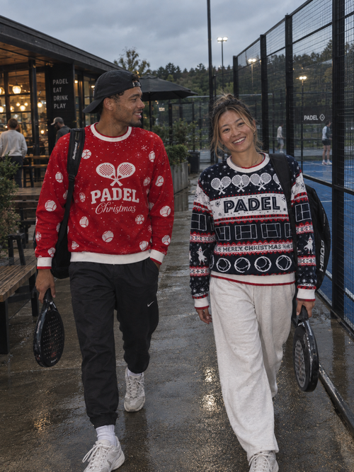 A man and a woman walking side-by-side on a wet paved path near a padelcourt, wearing festive Christmas sweaters. The man's sweater is red with white patterns and 'PADEL Christmas' text, and the woman's sweater is navy with red and white patterns and 'PADEL CHRISTMAS' text.
