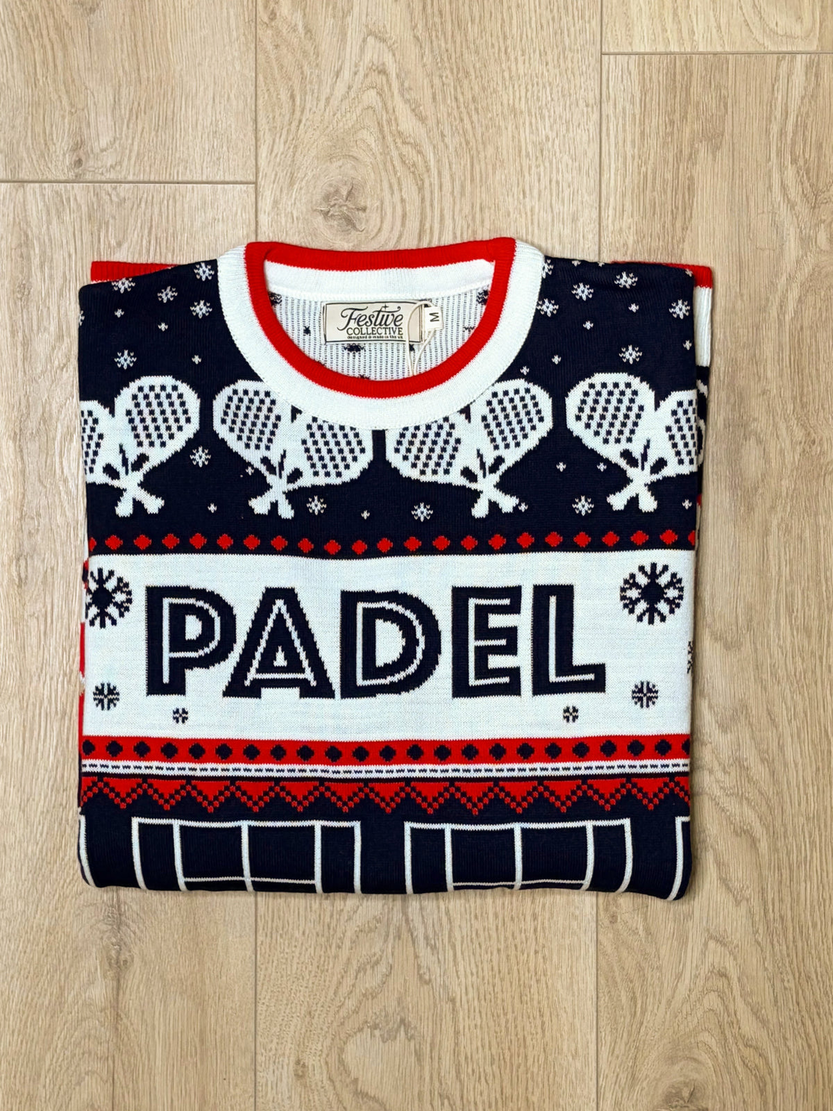 A folded navy blue knit sweater with white and red accents, featuring the word 'PADEL' across the front, laid flat on a light wood floor.